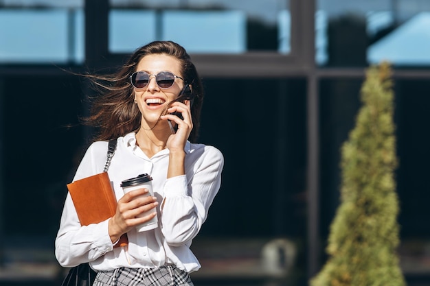 Donna d'affari che cammina e parla al cellulare all'aperto vicino al moderno centro affari con il taccuino