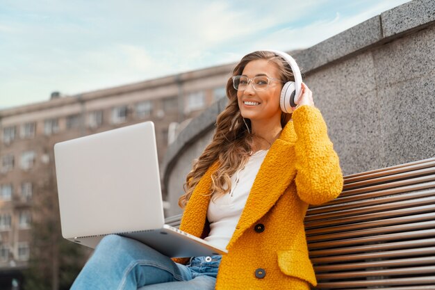 Business Woman Wear Eyeglasses Listen Music Headphone Outdoor Sitting Bench Using Laptop Outdoor Dressed Stylish Yellow Coat Smile Caucasian Female 30s Enjoy Podcast Or Audio Books Outside