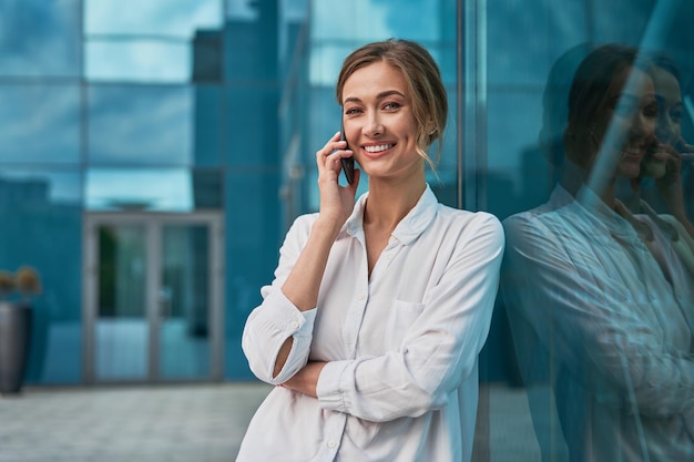 Business woman in a white blouse with a smartphone in her hands on the street, against the background of a business center. Work online. A serious expression on his face.