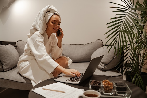 A business woman in a white robe and a towel uses a laptop and drinks tea or coffee. She is sitting on the sofa. Evening relaxation.