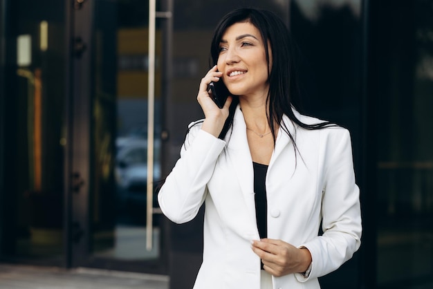 Деловая женщина в белом костюме разговаривает по телефону у офисного здания