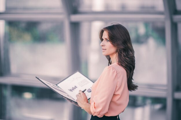 Деловая женщина с финансовыми документами стоит возле большого окна в офисе. На фотографии есть пустое место для вашего текста
