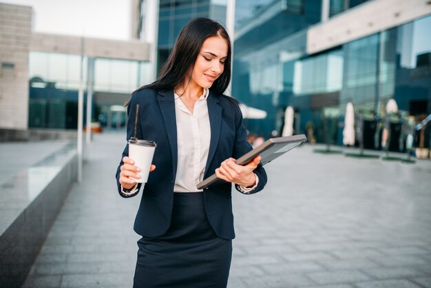 Business woman with laptop and coffee outdoor. Modern building, financial center, cityscape. Female businessperson in suit