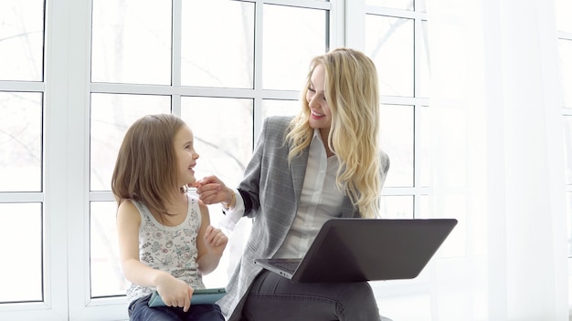 Business woman with laptop and little girl with tablet sitting by the big window.