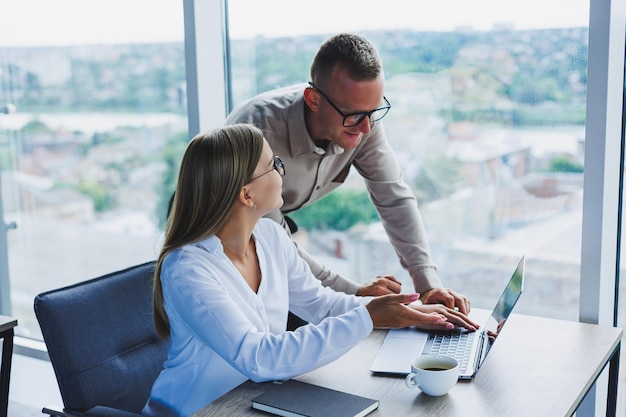 Business woman with a laptop for a male colleague who looks into an open notebook Business cooperation and teamwork European millennials at the table in the office Modern successful people