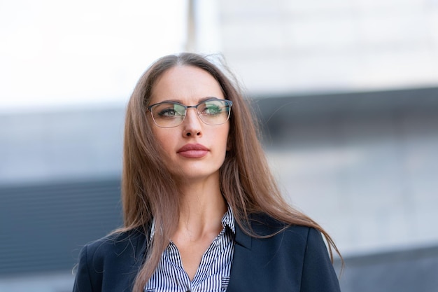 Business woman with long hair dressed black jacket eyeglasses standing outdoor near corporate office building close up Beautiful caucasian female business person portrait on city street Side view