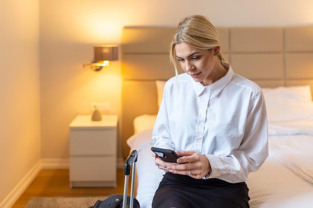 Business Woman with luggage in hotel room using her smart phone. Beautiful young woman holding her mobile phone. Sitting on the bed, business trip