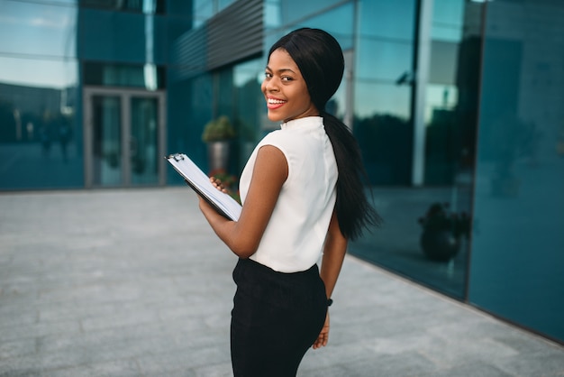 Photo business woman with notepad outdoors, back view, office building on background. black businesswoman in skirt and white blouse