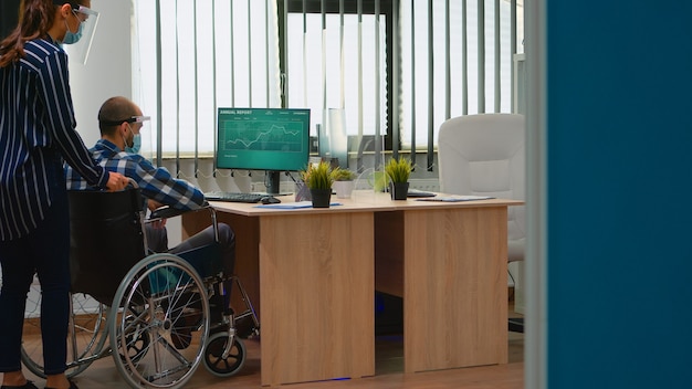 보호 마스크를 쓴 비즈니스 여성은 휠체어를 탄 장애인이 책상에 앉을 수 있도록 돕고, 코로나바이러스 전염병 동안 기업 사무실에서 새로운 정상으로 일합니다. 사회적 거리를 존중하는 기업인
