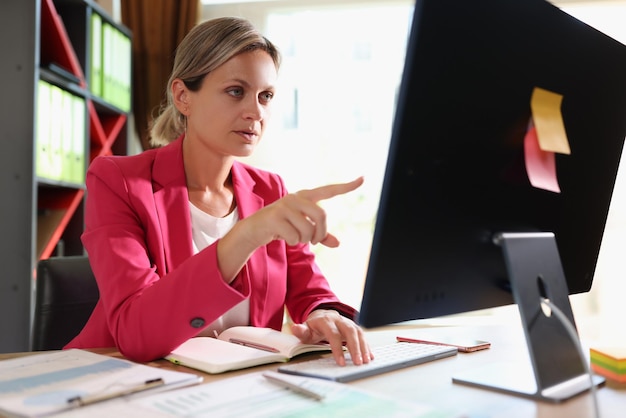 Деловая женщина с серьезным лицом за офисным столом указывает пальцем на экран компьютера