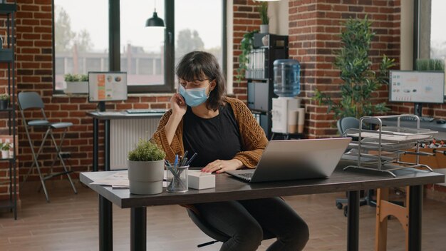 Photo business woman working with laptop and rate charts during covid 19 pandemic. startup employee using papers and computer for project planning and data analysis, wearing face mask.