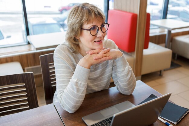 Business woman in the workplace at the window in the office A middleaged woman works on a laptop in a cafe She smiles