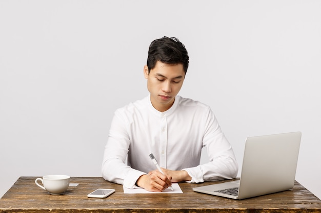 Business, work and interview concept. Busy serious handsome, young asian man sitting desk in office drink coffee, writing report, studying documents, use laptop, smartphone,