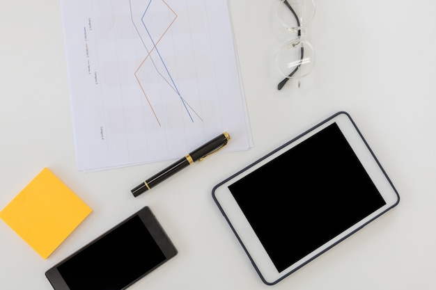 Business and Workplace concpet. Top view of work desk with blank screen tablet, smart mobile phone document paper line chart, pen notes and reading glasses with copy space.