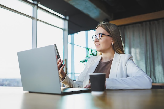 Giovane donna d'affari con gli occhiali e abiti casual seduti a un tavolo di legno con un computer portatile e una tazza di caffè che fa i compiti su un computer portatile