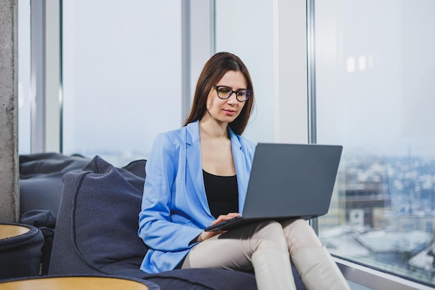 Деловая молодая женщина в очках с длинными темными волосами в повседневной одежде улыбается и смотрит на ноутбук, просматривая документы онлайн в выходные дни на рабочем месте