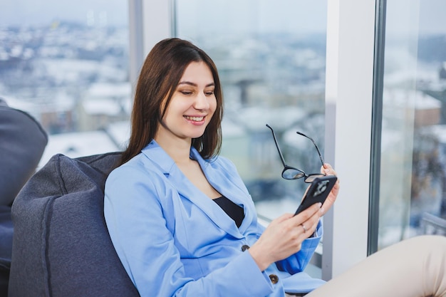 Деловая молодая женщина в очках с длинными темными волосами в повседневной одежде улыбается и смотрит на телефон, просматривая смартфон в выходной день на рабочем месте