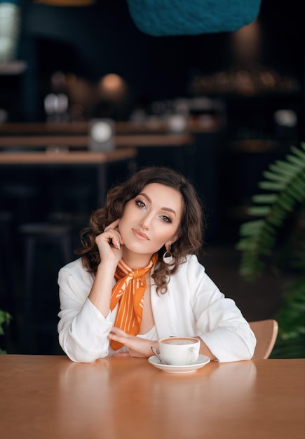 Фото Деловая молодая женщина сидит за столиком в кафе и пьет кофе, на ней белая куртка