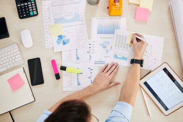 Businessman analyzing information in charts and diagrams and writing on sticky notes when preparing documents for colleagues, view from above