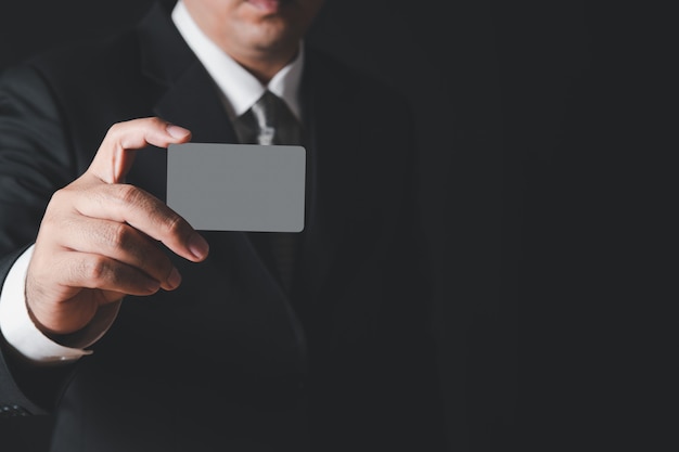 黒の衣装と黒の壁に灰色のカードを示す灰色のネクタイのビジネスマン。ペイメントバンキングのコンセプト