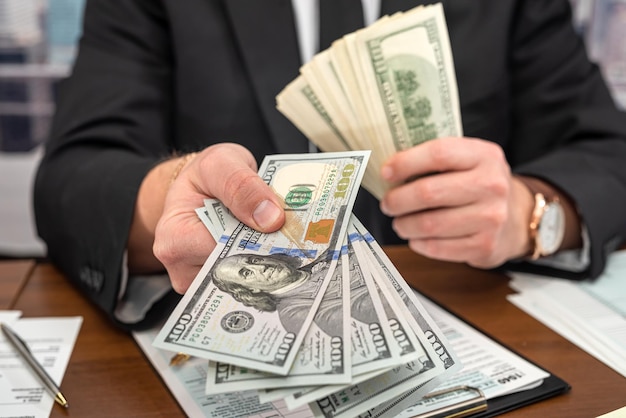 Businessman in a black stylish suit at a spacious table counts\
dollars and fills out forms