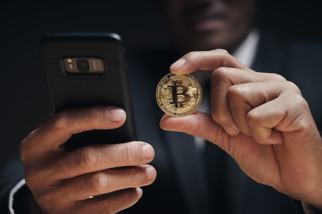 businessman in black suit holding a golden bitcoin using smartphone with bitcoin trading chart on the screen to trading cryptocurrency, alternative finance and investment concept.