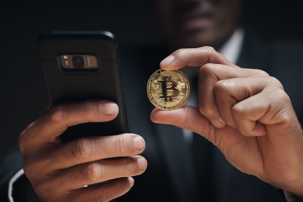 Businessman in black suit holding a golden bitcoin using Smartphone with Bitcoin trading chart on the screen to Trading Cryptocurrency, alternative finance and investment Concept.