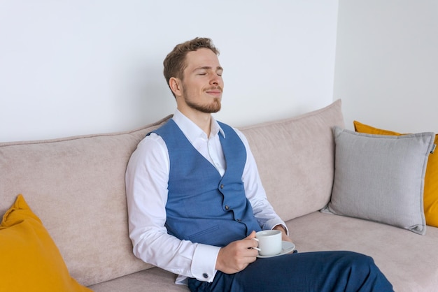 彼のオフィスのソファに座っている青いスーツのビジネスマンはコーヒーカップと一緒に座っています