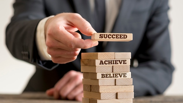 Businessman building tower of wooden domino bricks with motivational concept signs Believe, Achieve and Succeed.