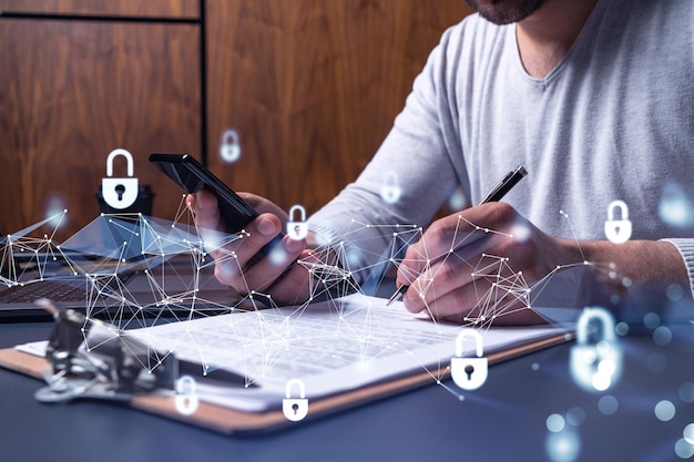 A businessman in casual wear signing the contract to prevent\
probability of risks in cyber security checking the phone padlock\
hologram icons over the working desk