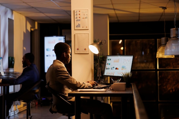 Businessman checking marketing report on computer, analyzing financial graphs while working overhours at company strategy. African american manager planning management research plan