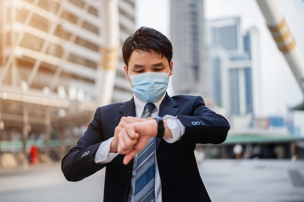 그의 시계에서 시간을 확인하고 도시에서 코로나 바이러스 전염병 동안 의료 마스크를 착용하는 사업가