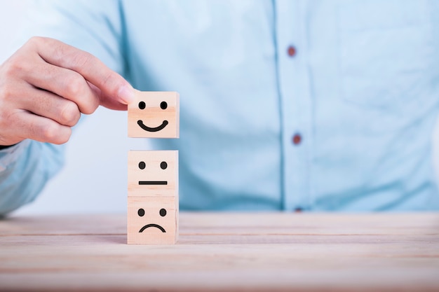ビジネスマンは、木製のブロック、サービスおよび顧客満足度調査の概念に笑顔の絵文字アイコン顔幸せなシンボルを選択します