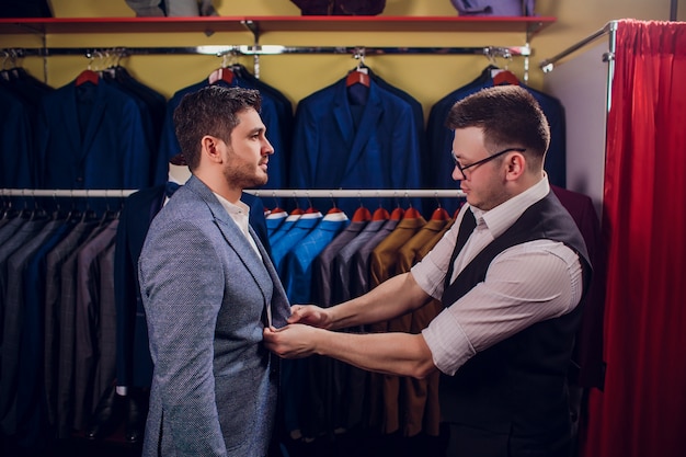 Businessman in classic vest against row suits in shop. Man helps another try on suit in clothing store