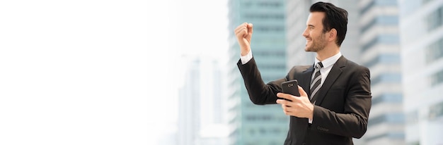 Photo businessman clenching fist while holding mobile phone against building