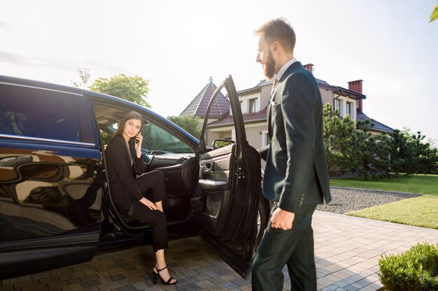 Photo businessman coming to car and opens door for young woman, talking on the phone. businesswoman get out from the auto and is ready to go with her colleague to office building