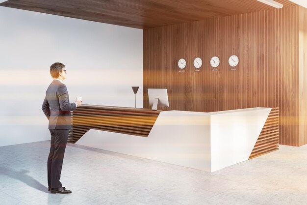 Foto uomo d'affari nell'angolo dell'ufficio moderno con pareti bianche e in legno, pavimento piastrellato e reception bianca e in legno con computer e orologi sulla parete. immagine tonica