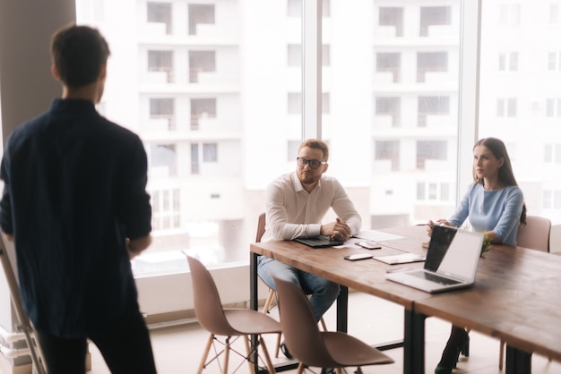 Businessman doing presentation using whiteboard explaining to\
colleagues in modern office meeting against large panoramic window\
concept of office life