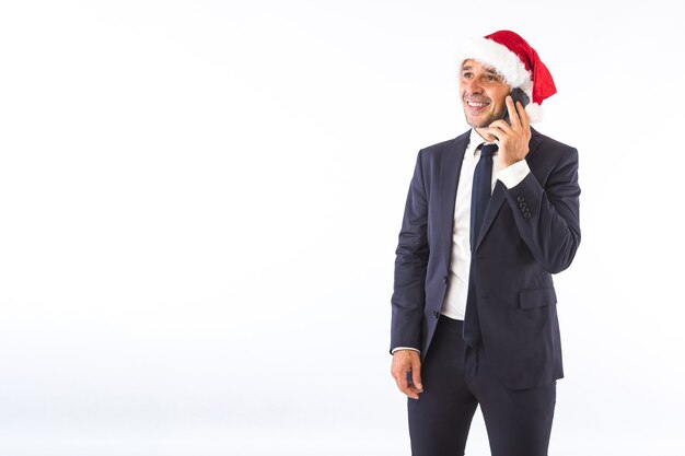 Businessman dressed in suit, tie and Christmas hat of Santa Claus, talking on the phone, on white background. Christmas celebration concept.