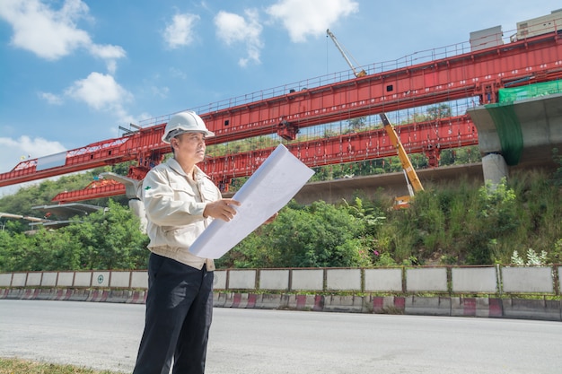 L'uomo d'affari o l'ingegnere o l'architetto che indossa l'elmetto protettivo e la maglia di sicurezza sorvegliano il progetto dell'autostrada o dell'autostrada