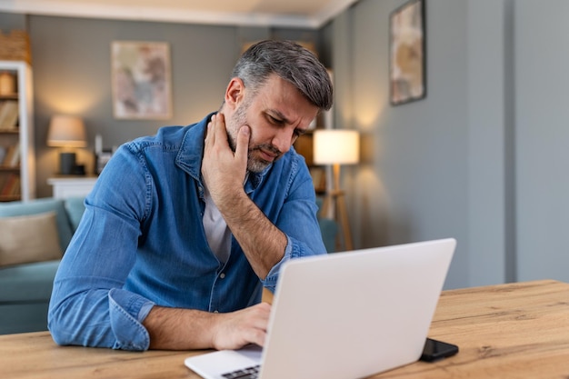 Businessman feeling pain in neck after sitting at the table with laptop Tired man suffering of office syndrome because of long hours computer work He is massaging his tense neck muscles