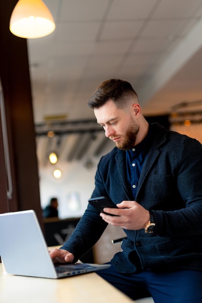 Businessman in formal clothes sits in cafe with laptop and phone in hands. Handsome and stylish young manager. Closeup.