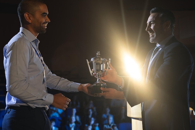 Photo businessman giving trophy to business male executive on stage in auditorium