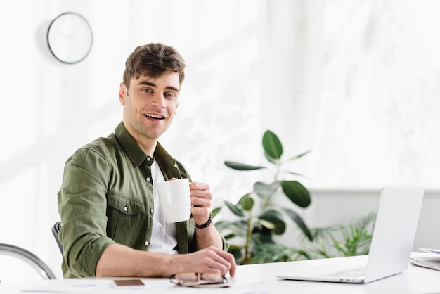 Businessman in green shirt sitting with cup at table with laptop in office