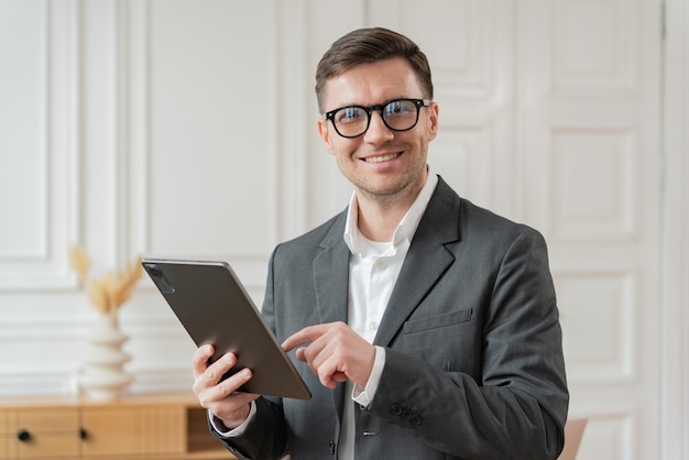 A businessman in a grey suit and glasses smiles while using a tablet in a sophisticated white office