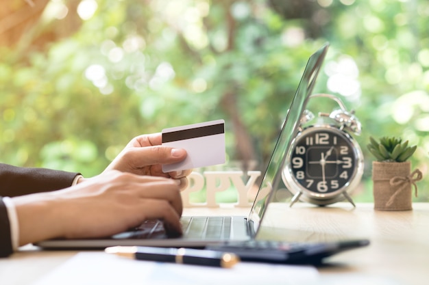 Businessman hand holding credit card and using laptop on desk with document and calculator.