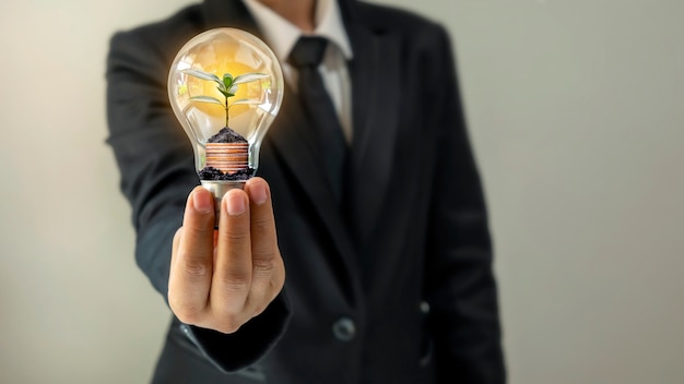 Businessman hand holding energy-saving light bulb with plants growing on coins in light bulbs