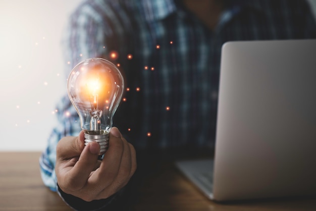 Lampadina della tenuta della mano dell'uomo d'affari con l'idea d'ardore e creativa arancio al computer portatile.