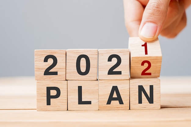 사업가가 나무 큐브를 들고 테이블 배경에서 블록 2021에서 2022 계획을 뒤집습니다. 해상도, 목표, 검토, 변경, 시작 및 새해 휴일 개념