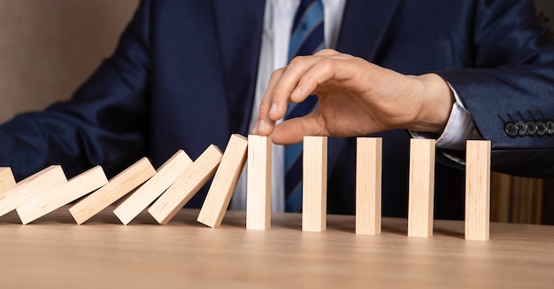 Businessman hand stopping falling wooden dominoes effect from\
continuous toppled or risk, strategy and successful intervention\
concept for business.